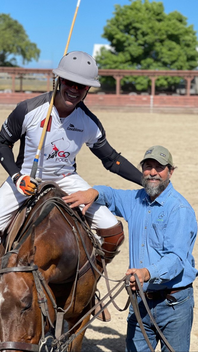 Daniel Fernández, in an Aluminé cap and shirt, laughs with a smiling rider on a bay horse after a ride.